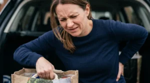 Woman stopped mid-lift at the boot of her car, one hand pressed into her lower back after straining it lifting shopping